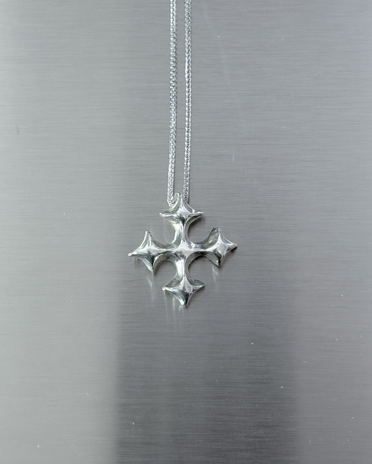 A silver cross pendant hanging from a delicate silver chain against a brushed metal background.
