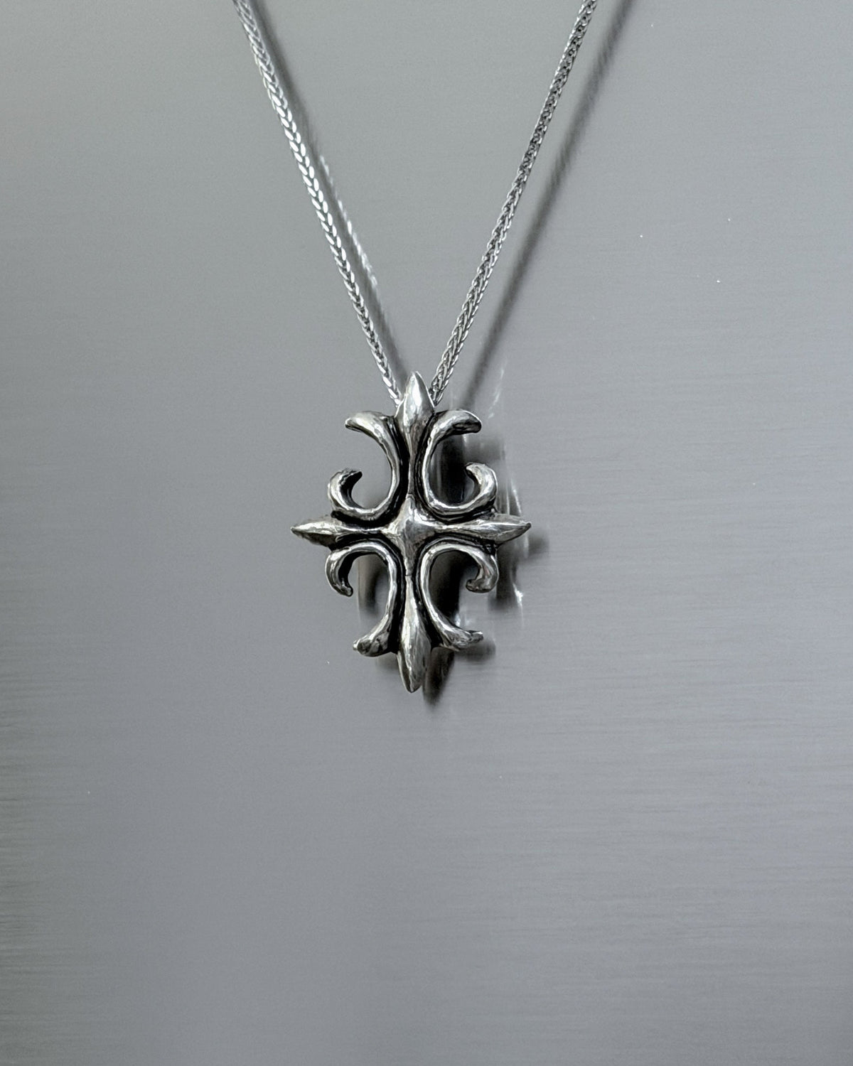 A silver cross pendant necklace with an ornate, textured design hanging from a delicate silver chain against a brushed metal background.