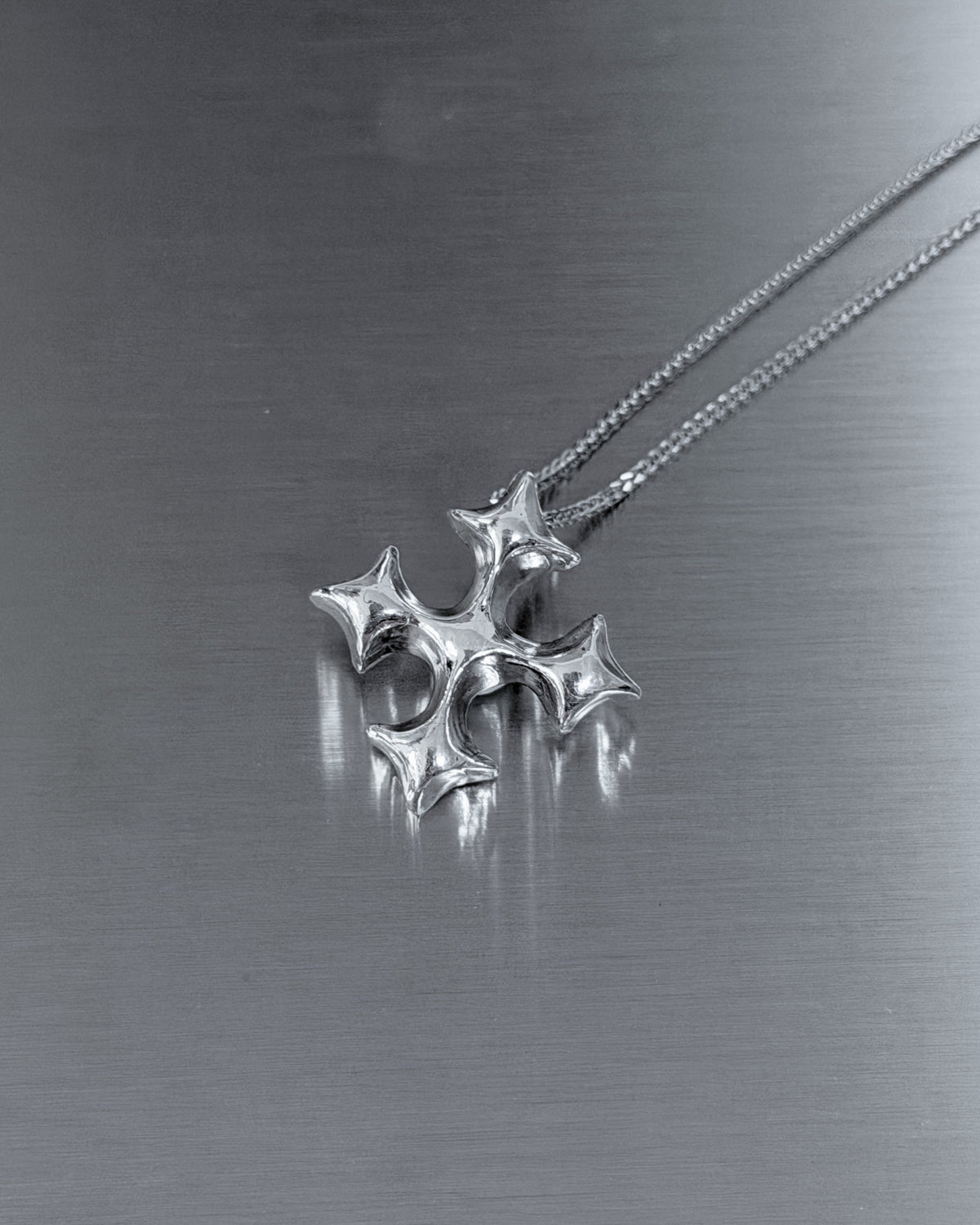 A silver-toned metal pendant necklace with a four-pointed star or cross-like design hanging from a fine chain on a brushed metallic gray surface.