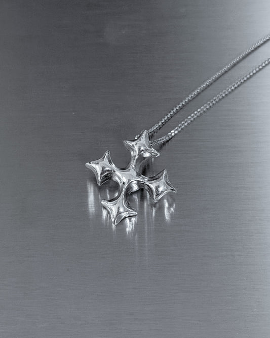 A silver-toned metal pendant necklace with a four-pointed star or cross-like design hanging from a fine chain on a brushed metallic gray surface.