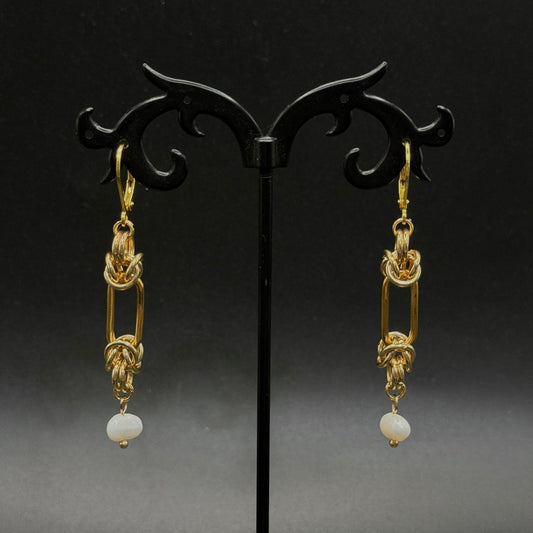 Gold stainless steel earrings with pearl drops on a black stand against a dark background