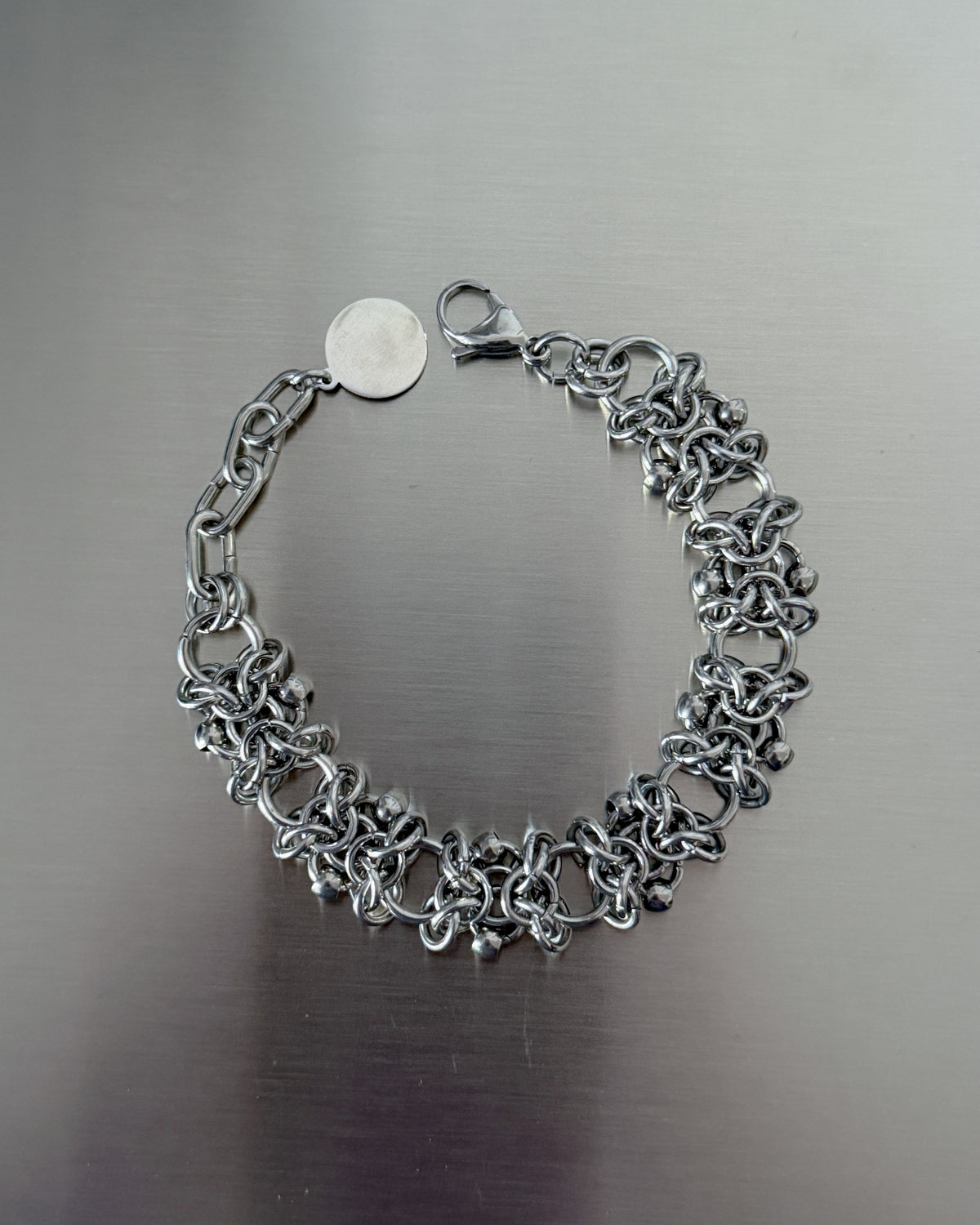 A silver-toned chain link bracelet with a lobster clasp and a small round tag, displayed on a brushed metal surface.