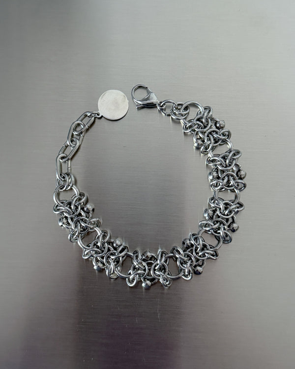 A silver-toned chain link bracelet with a lobster clasp and a small round tag, displayed on a brushed metal surface.