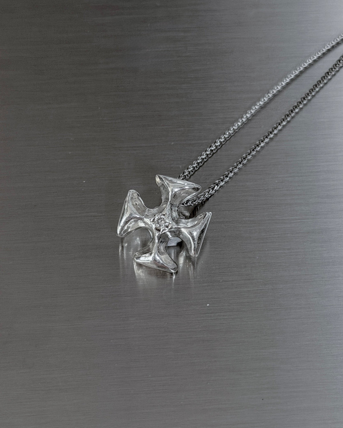 A silver necklace with a detailed cross pendant resting on a brushed metal surface.