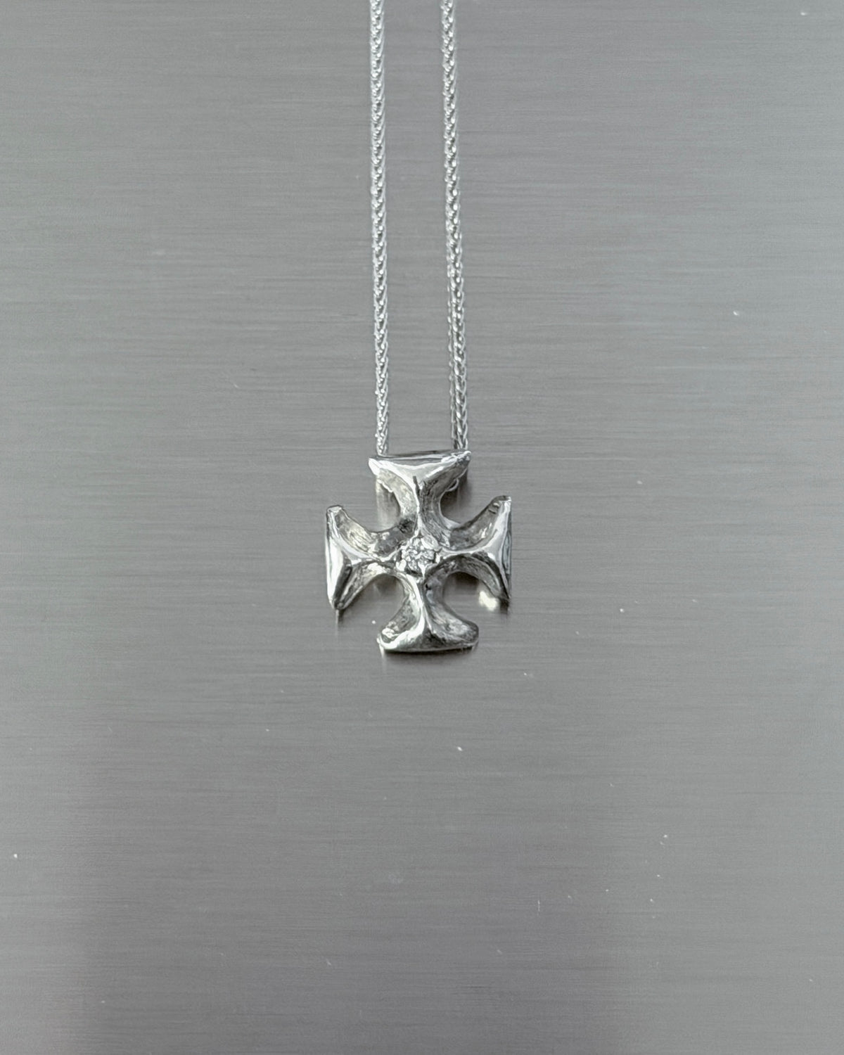 A silver cross pendant necklace with a textured finish and a small clear stone in the center, hanging from a delicate silver chain against a brushed gray metal background.