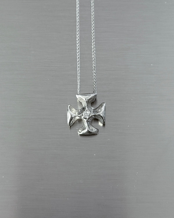 A silver cross pendant necklace with a textured finish and a small clear stone in the center, hanging from a delicate silver chain against a brushed gray metal background.