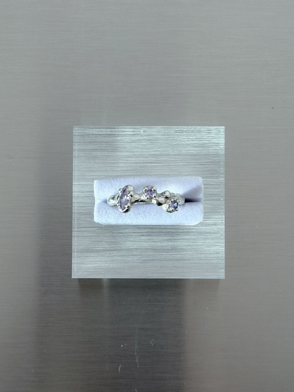 A silver ring with three small lavender gemstones displayed on a white insert inside a clear acrylic block, set against a brushed gray metal background.