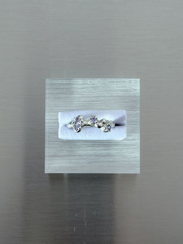 A silver ring with three small lavender gemstones displayed on a white insert inside a clear acrylic block, set against a brushed gray metal background.