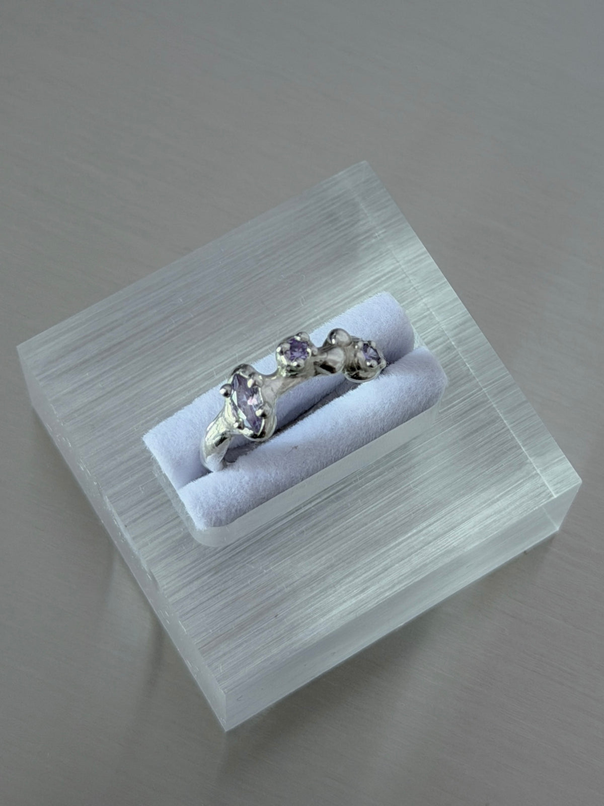 A silver ring with three small purple gemstones displayed on a clear acrylic ring box with a white velvet cushion, set against a neutral gray background.