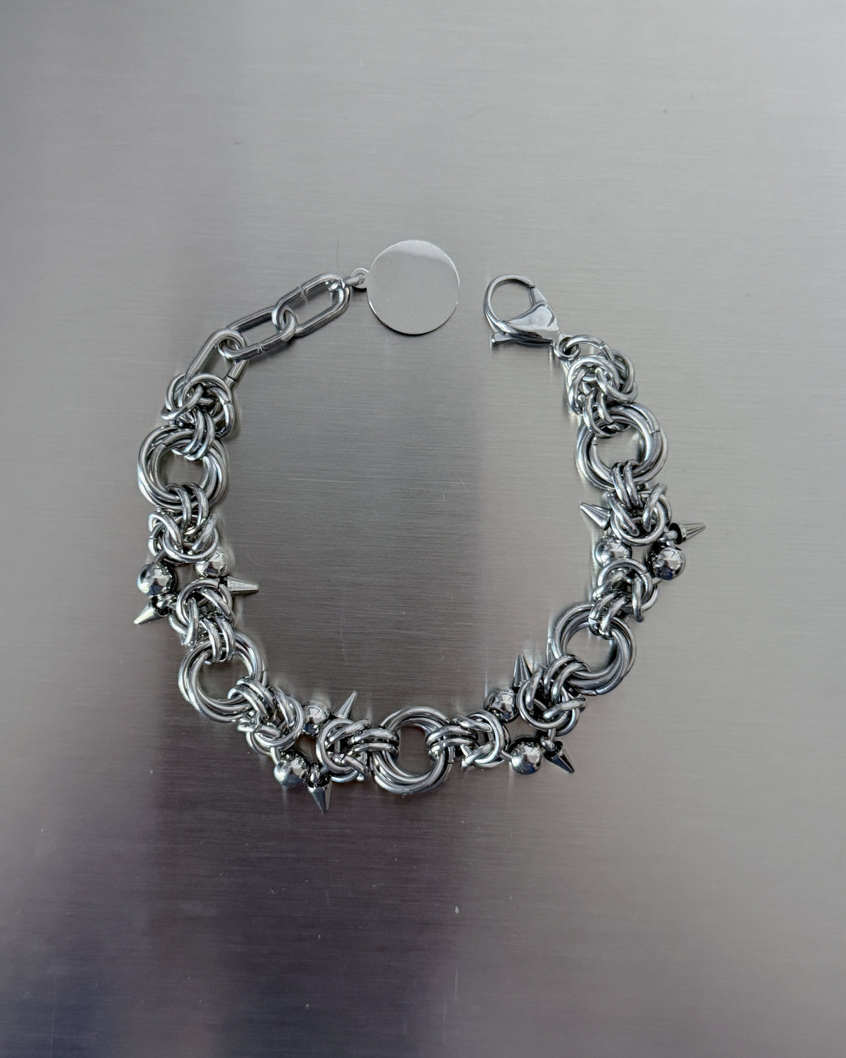 A silver-toned metal chain bracelet with small spiked and ball-like charms, displayed on a brushed metallic surface.
