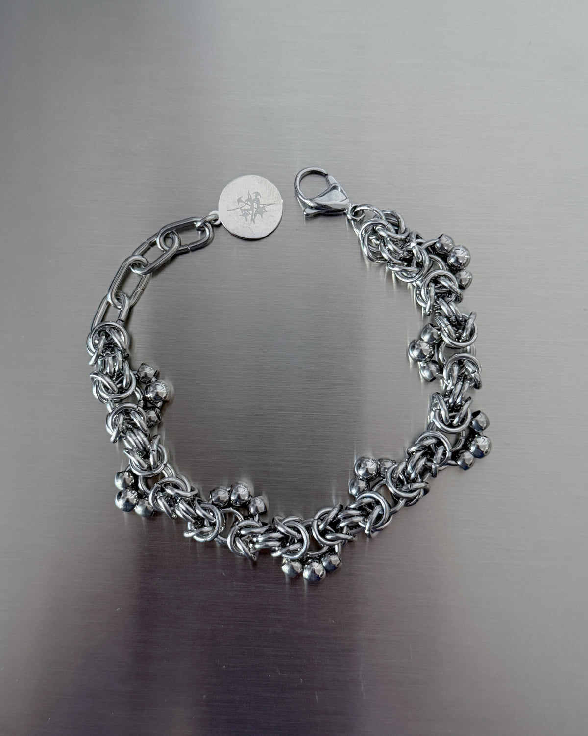 A silver-toned chain bracelet with a lobster clasp and a round engraved tag, displayed on a brushed metal surface.