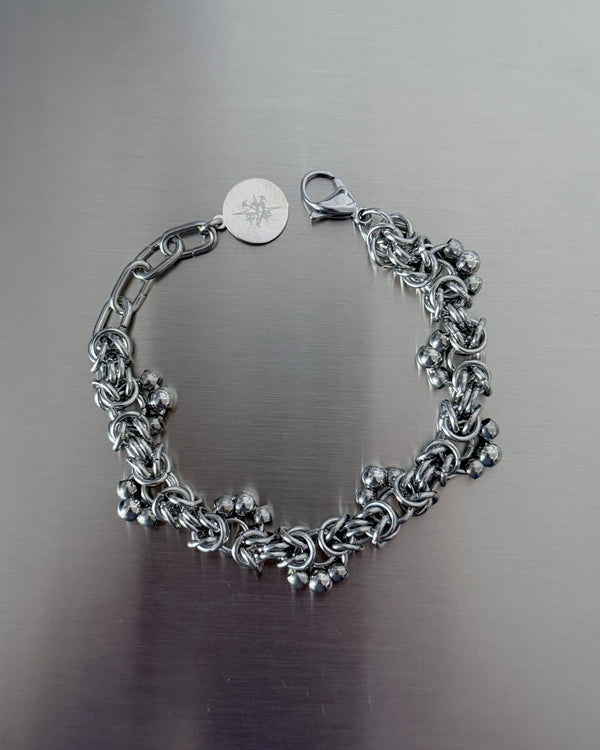 A silver-toned chain bracelet with a lobster clasp and a round engraved tag, displayed on a brushed metal surface.