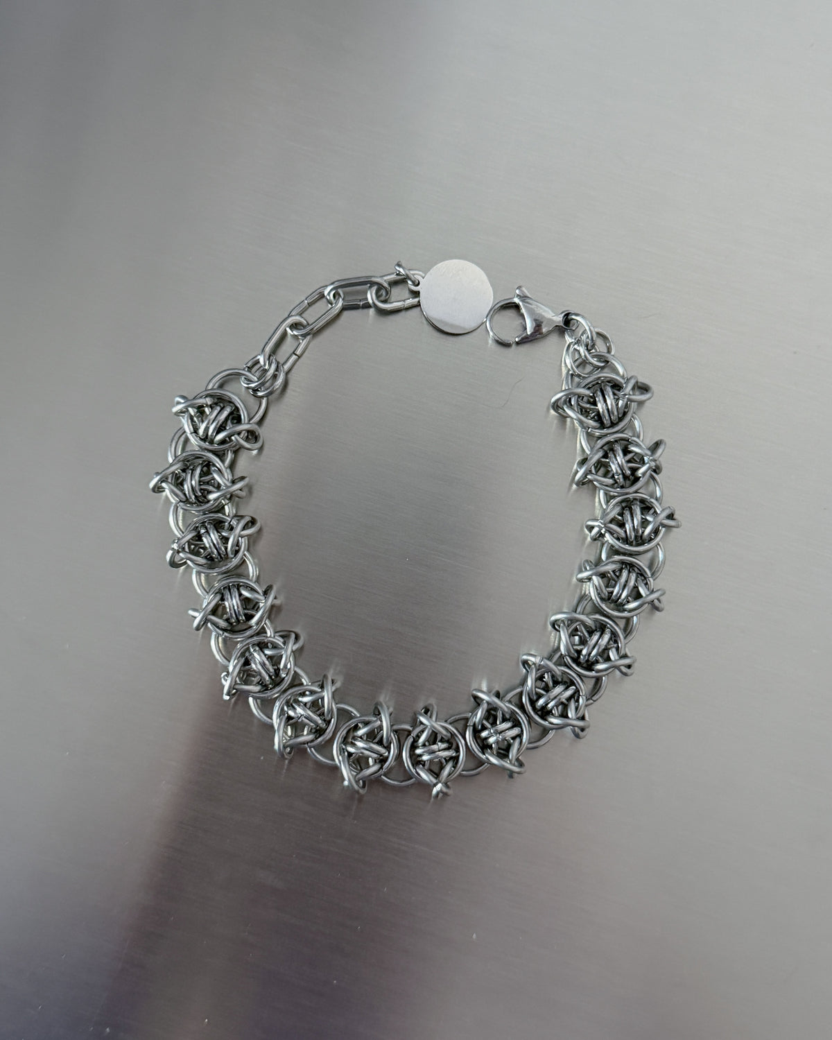 A silver-toned metal bracelet with a textured, interlocking link design and a round white tag, displayed on a brushed gray metal surface.