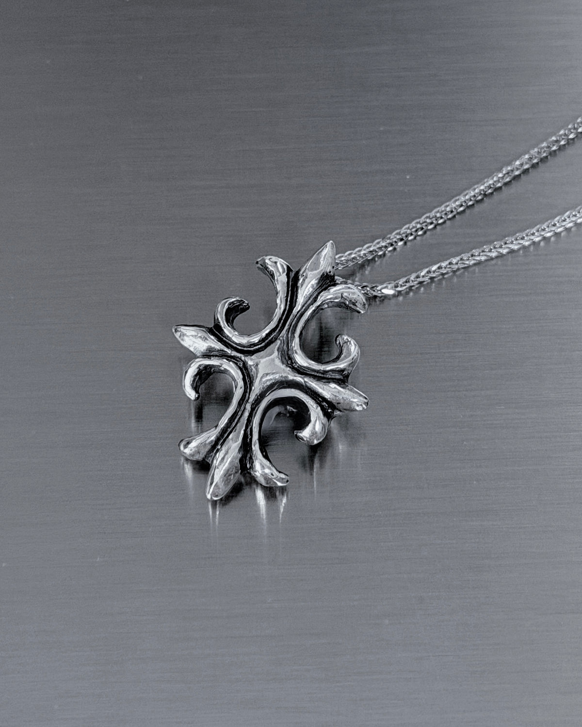 A silver-toned metal pendant necklace with an ornate, stylized cross-like design, resting on a brushed dark gray metal surface.