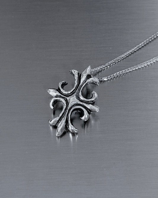A silver-toned metal pendant necklace with an ornate, stylized cross-like design, resting on a brushed dark gray metal surface.