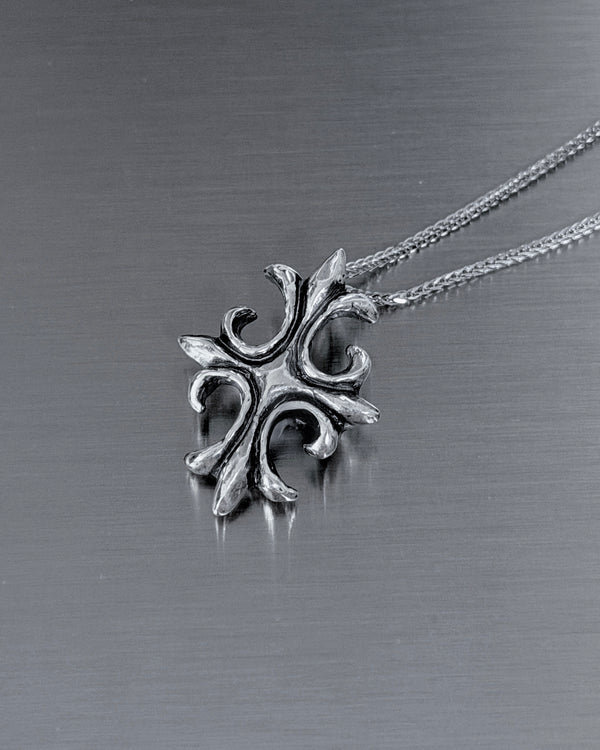 A silver-toned metal pendant necklace with an ornate, stylized cross-like design, resting on a brushed dark gray metal surface.