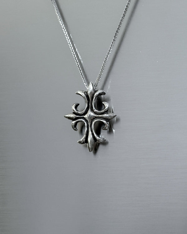 A silver cross pendant necklace with an ornate, textured design hanging from a delicate silver chain against a brushed metal background.